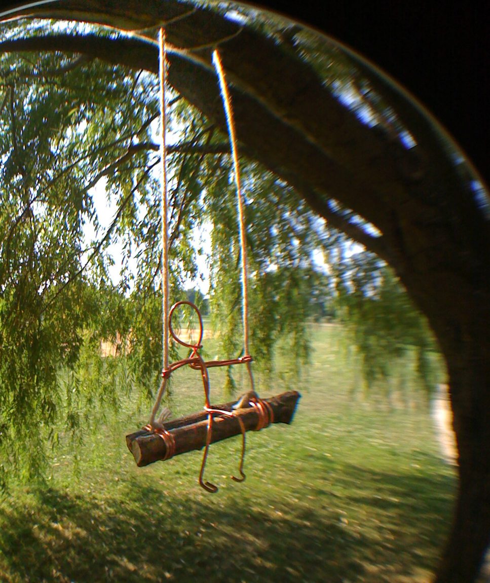 A vertical, close-up photograph of a miniature, handmade art installation hanging from a tree branch in a sunlit park. The image is captured through a circular glass lens or prism, creating a soft, blurred vignette around the edges that centers the focus on a small wire figure.
The figure is minimalist, crafted from single strands of copper-colored wire. It sits on a swing made of a thick, dark wooden stick found on-site, which is suspended by two thin white strings tied to a branch above. The wire person is depicted in a relaxed posture: its circular head is tilted slightly upward, its thin wire arms reach out to hold the string supports, and its legs are bent at the knees, dangling peacefully over the edge of the wooden seat.
The background, seen through the weeping branches of a willow tree, reveals a lush green field and a distant baseball diamond under a bright, clear sky. The sunlight filters through the willow leaves, casting a warm, dappled glow over the wire figure and the grass below. The overall composition emphasizes a sense of scale, playfulness, and quiet observation.