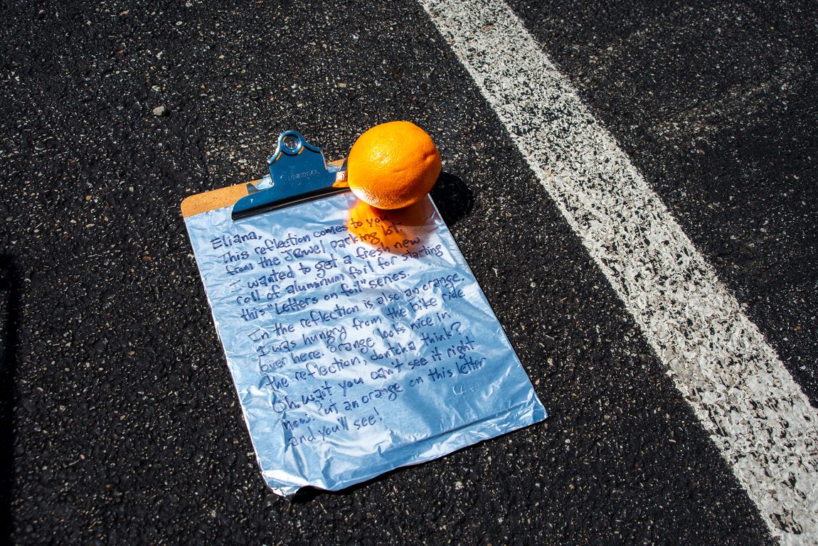 Bright orange sits on a clipboard with aluminum foil. On the foil is a letter. The clipboard is on an asphalt parking lot for a suburban grocery store.