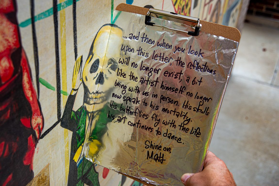 Clipboard wrapped in aluminum foil is positioned against the Tony Fitzpatrick mural in Glen Ellyn, Illinois. The foil reflects a dancing figure with a skull head. The aluminum foil has a letter written with a Sharpie marker. The letter says, ".. and then when you look upon this letter, the reflecctions will no longer exist, a bit like the artist himself no longer being with us in person. His skulls now speak to his mortality. His butterflies fly with the life his art continues to dance."