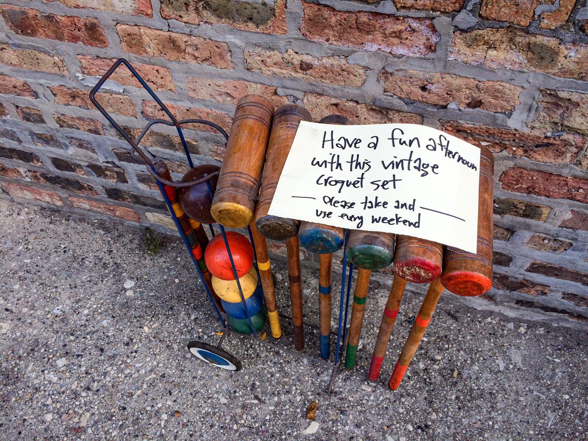 Vintage croquet set in wire rack with wheels. Sitting in Chicago alley by chicago brick wall. Handwritten sign on croquet set says, “Have a fun afternoon with this vintage croquet set. Please take and use every weekend.”