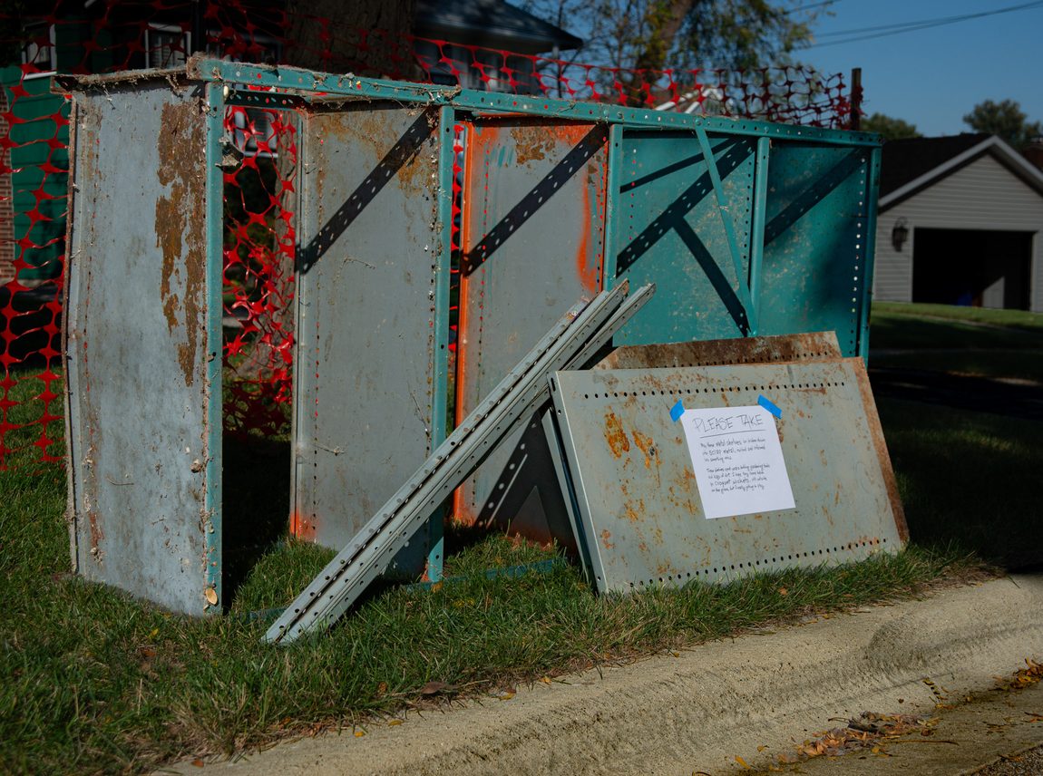 Old metal shelf painted teal is laying on its side by the curb in suburban Glen Ellyn of Chicago, Illinois. A second shelf (gray color) is disassembled into parts leaning up against the teal shelf. The parts have a letter-sized sign with a handwritten message that says, "PLEASE TAKE. May these metal shelves be broken down into scrap metal, melted and reformed into something new. These shelves spent years holding garden tools in the dirt. I hope they come back as croquet wickets, still outside on the grass, but finally getting to play."
